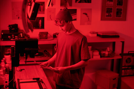 Waist up portrait of teenage boy looking at developed photo prints in darkroom lit by red light young photographer in creative studio copy space