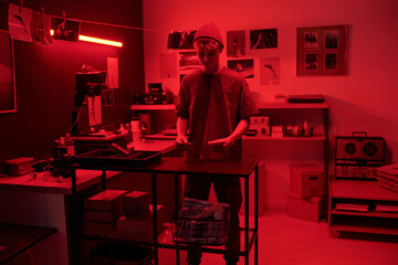 Full length portrait of young man as photographer working in darkroom lit by red light and developing analog photos in creative studio copy space