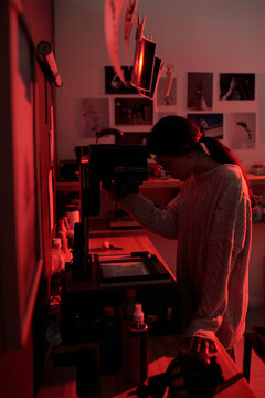 Artistic side view portrait of teenage girl working with enlarger while developing analog photos in darkroom lit by red light young photographer in creative studio