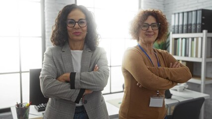 Confident hispanic women partners in an office setting stand with arms crossed, reflecting teamwork and professionalism amid modern, indoor workspace environment.