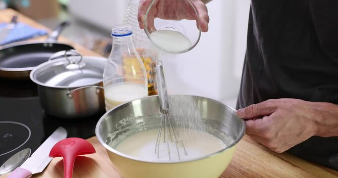 Man adds sugar to liquid crepe dough in metal bowl on table. Male cooks homemade pancakes of natural ingredients in light home kitchen slow motion
