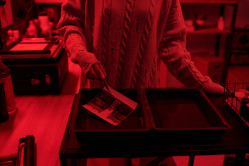 Close up of young woman dipping developed photo print in chemical tray while working on analog prints in darkroom lit by red light copy space