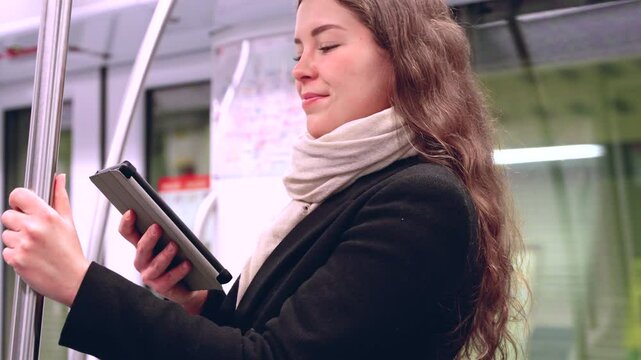 Stylish young woman in a black coat and beige scarf reading on an e-reader while standing inside a modern subway train. She balances while holding onto a pole during her commute. 