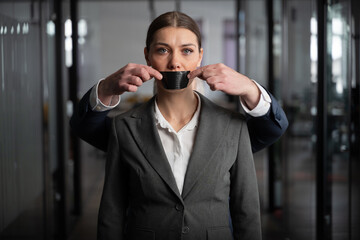 Woman in a suit stands in an office with a serious expression, as a man behind her silences her with black tape over her mouth. Workplace oppression, censorship, and gender inequality