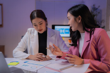 Two Asian businesswomen discussing marketing strategies while working late at night in an office, analyzing financial graphs with a laptop and pen, showcasing collaboration and teamwork