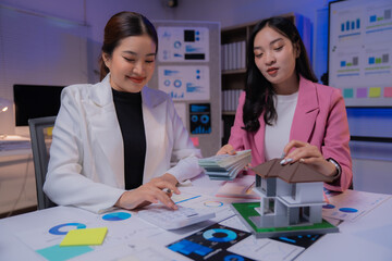 Two businesswomen working together at modern office desk at night, discussing housing project details, investment and financial income, using calculator and holding house model and dollar banknotes