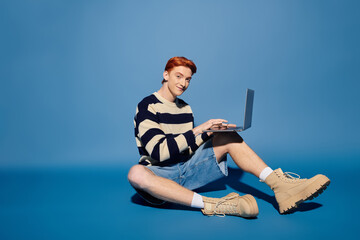Emotional young man engaged in creative activities on a laptop against a blue backdrop