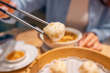Metal chopsticks holding a xiaolongbao dumpling over a bamboo steamer basket in a restaurant