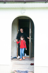 A young girl with pigtails exits a house with a woman. The girl is wearing a red shirt, jeans, and...