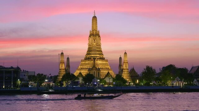 Long boat passing on river in front of illuminated Wat Arun at sunset