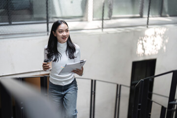 Young professional woman walking up stairs with coffee and documents, smiling in a modern office environment.