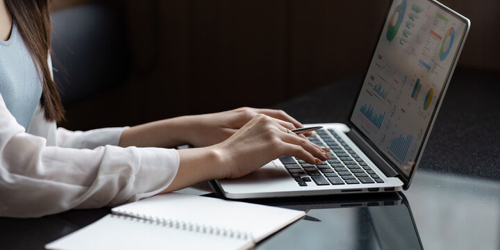 Professional Asian woman analyzing data on a laptop while taking notes in a modern office.