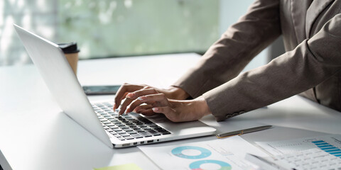 Diverse business professional typing on a laptop while analyzing financial data and charts in a modern office setting.