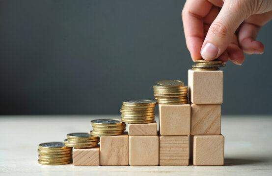 Hand puts coins on wooden cube stairs. Growth of savings, financial planning, investment, pension contribution. Long term future vision, capital rise, retirement savings, wealth accumulation,