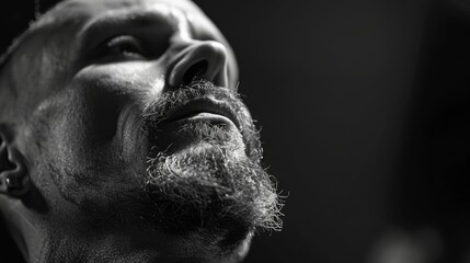 A man with a well groomed beard and intense expression looks upward, illuminated by soft light, highlighting his facial features in a moody atmosphere.