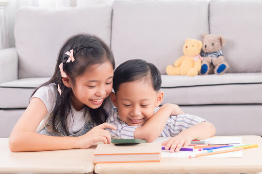 Asian siblings laughing together while using digital tablet. Young boy and girl sitting at table watch cartoons at home. Bright indoor setting, joyful expression, sibling bonding, technology use.