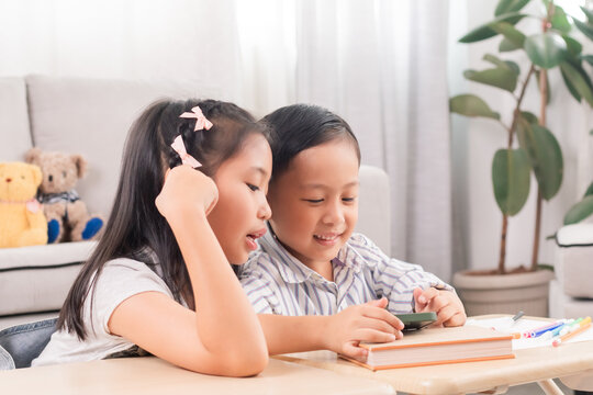 Asian siblings laughing together while using digital tablet. Young boy and girl sitting at table watch cartoons at home. Bright indoor setting, joyful expression, sibling bonding, technology use.