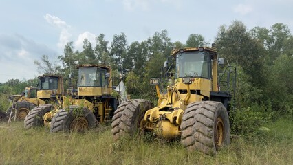 Obraz premium Heavy yellow construction graders parked in a grassy field with lush green trees under a cloudy sky