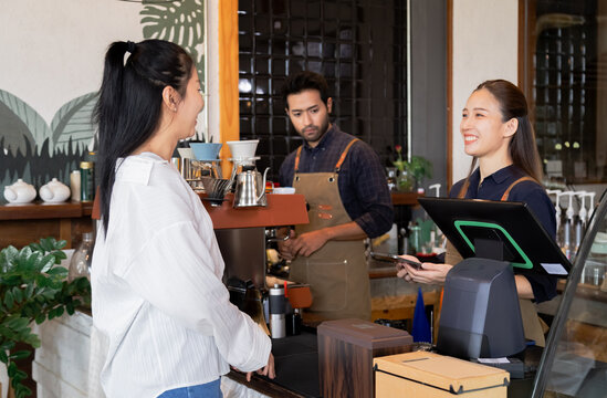 Asian cafe cashier worker female showing menu or cashless billing payment to customer, barista small business owner using technology tablet receives coffee order at workplace in restaurant coffee shop