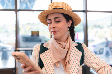 Portrait of young pretty Caucasian woman wearing straw hat using smartphone. Digital app for trip. Concept of vacation and travel