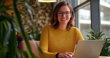 Une femme heureuse portant des lunettes, assise à une table avec un ordinateur portable et souriant à la caméra, vêtue d'un pull jaune.
