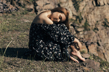 A woman sits on the ground, leaning on her knees and closing her eyes. She is wearing a dark dress with a floral pattern. Her hair is light brown and falls loosely over her shoulders.