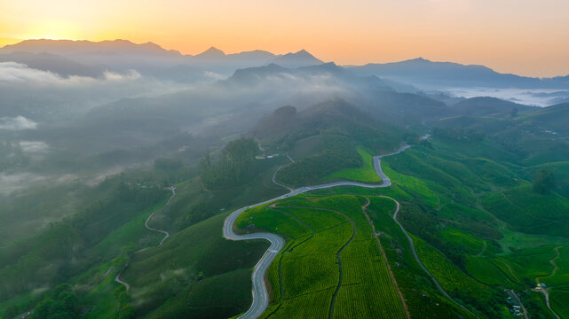 A stunning drone shot of the Gap Road in Munnar, winding through lush green tea estates at sunrise. The golden light highlights the misty landscape, with majestic mountains in the background, creating