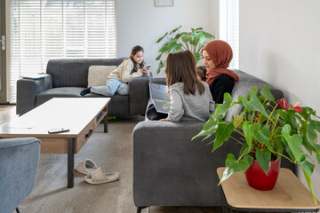 Mother wearing a hijab sits with two daughters on a sofa, both focused on a smartphone, while another sister lounges with a smartphone on a separate couch in a bright living room, Netherlands