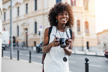 Young black female tourist enjoys walking through the streets of a beautiful European city. She is happy and using her photo camera to take fantastic architecture photographs. Bright sunny day. © Dusko