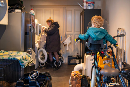 A young child on a bicycle observes mother in a coat exiting a cluttered home interior, featuring a laundry area and various household items, Netherlands