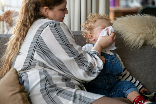 A mother wipes her young child's nose with a tissue while sitting on a sofa in a cozy home setting, Netherlands