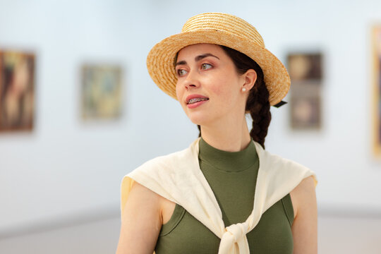 Portrait of a young Caucasian elegant smiling woman in straw hat posing in the art gallery. Paintings in background. Concept of cultural education and Museum Day