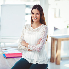 Smiling businesswoman in office leaning on desk with arms crossed confidently