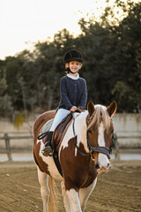 A young girl rider in a helmet and equestrian boots sits on her beloved horse at the stable,...