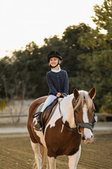 A young girl rider in a helmet and equestrian boots sits on her beloved horse at the stable,...
