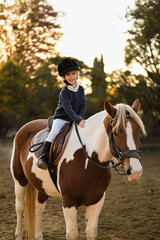 Obraz premium A young girl rider in a helmet and equestrian boots sits on her beloved horse at the stable, enjoying a peaceful horseback ride.