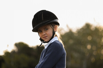 A young girl rider in a helmet and equestrian boots sits on her beloved horse at the stable,...