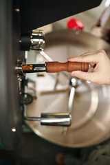 Vertical high angle shot of unrecognizable male hand taking scoop with coffee beans from professional machine to check roasting level