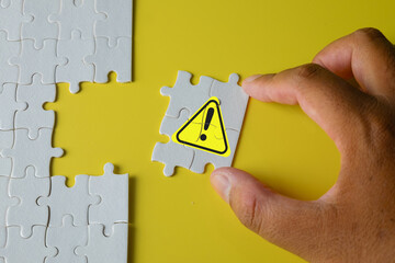 A hand inserting a final puzzle piece with a yellow warning sign, representing business risk,...