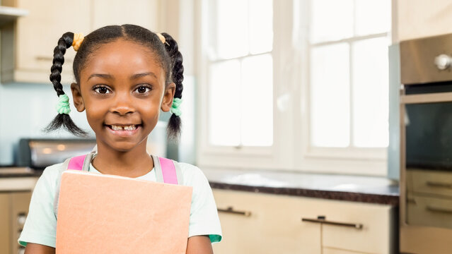Smiling African American girl holding book in kitchen, ready for school, copy space