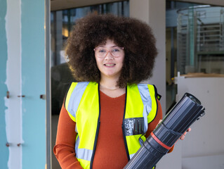 Holding blueprint tube, engineer in high-visibility vest smiling confidently at office