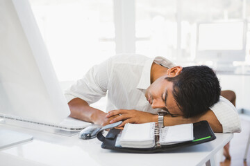 Exhausted businessman sleeping at desk with planner and computer in office