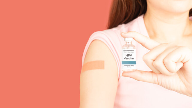International HPV awareness day concept. Woman holding HPV vaccine vial while showing vaccinated arm with medical adhesive bandage strip after vaccine injection over pink background.