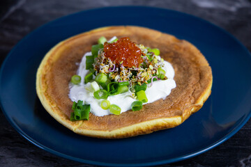 A delicious golden pancake topped with sour cream, fresh greens, and red caviar, served on a blue...