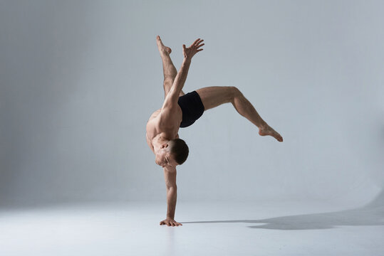 Professional male dancer in black shorts balances on one hand in an acrobatic pose in a studio with white background, displaying strength and grace.