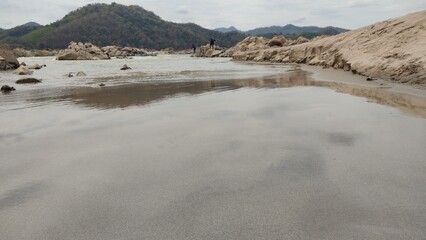 Rocky dry riverbed landscape with scattered stones and boulders under a partly cloudy sky. Mountainous background with green hills, depicting drought, climate change, and natural erosion effects