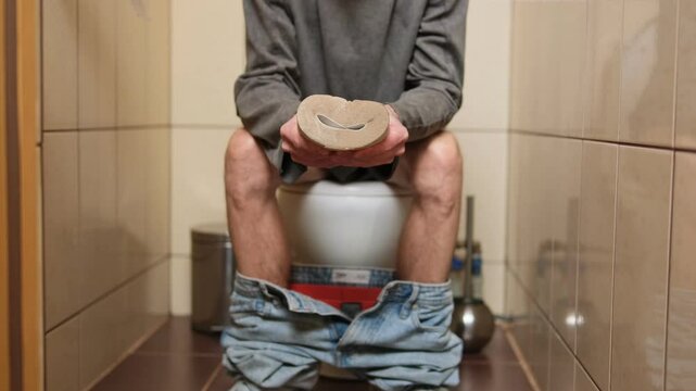 Man sitting on a toilet with pants down, tightly squeezing an empty toilet paper roll, expressing abdominal cramps. Symbolizing constipation, diarrhea, or digestive issues in a tiled bathroom.