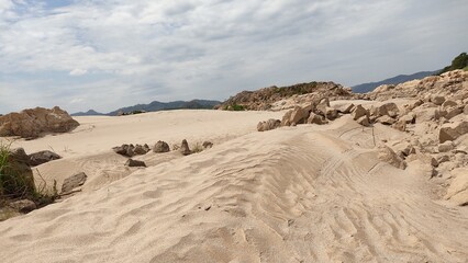 Shallow river water reflecting the cloudy sky, with fine sand patterns in the foreground. Rocky riverbanks and distant mountains create a serene natural landscape, perfect for travel, nature, and envi