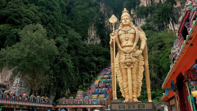 Golden murugan statue greeting visitors near limestone temple entrance at famous malaysian hindu pilgrimage site