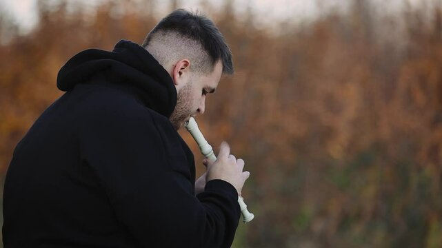 A young man in a black sweatshirt plays on panpipe enchanting melodies in the autumn park